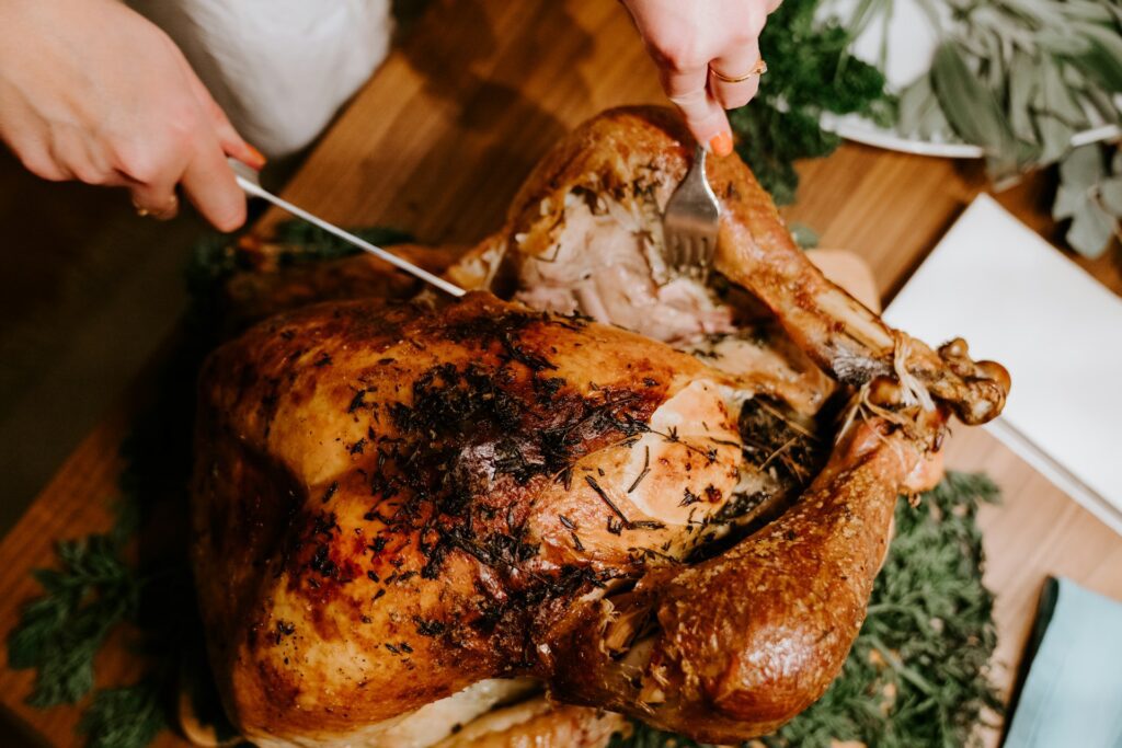 Hands carving a golden-brown, herb-roasted Thanksgiving turkey on a wooden cutting board garnished with fresh greenery.